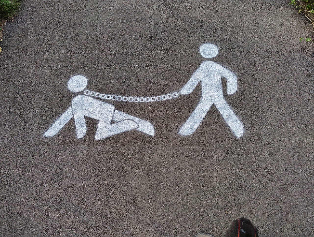 Logo peint au sol, inspiré des symboles de passage piéton (un bonhomme blanc stylisé), mais avec une particularité : le personnage tient en laisse (ou plutôt enchaîné) un second bonhomme à quatre pattes (comme s'il promenait un chien en laisse).